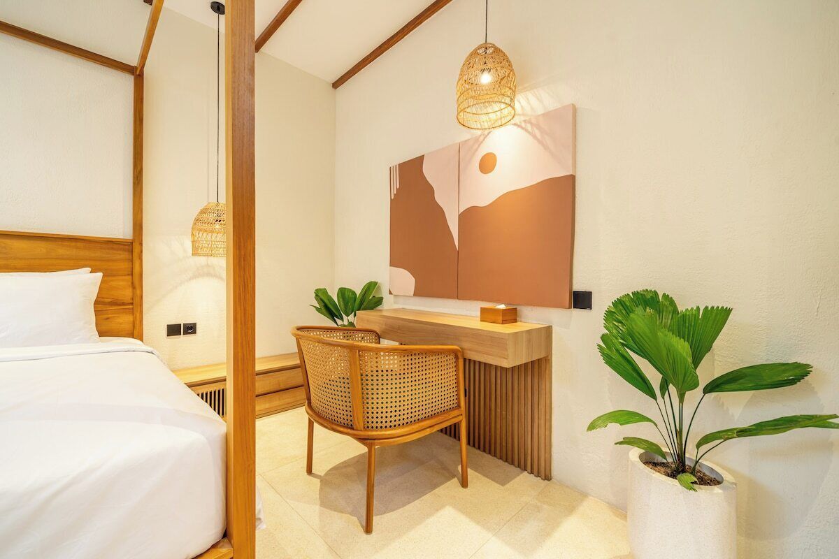 A hotel room with a bed, a desk, a chair, a hanging light fixture, and a potted plant.