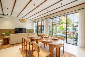 A modern dining area with a wooden table set for six, a sofa, and a television in the background. - Designer 3BR Villa in Ubud w Pool, Patio & Garden (Gianyar Regency)
