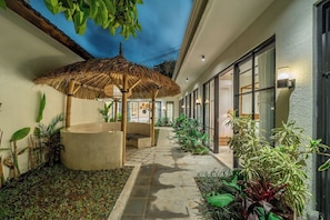 A covered outdoor seating area with a thatched umbrella, surrounded by potted plants and a stone pathway leading to a building entrance. - Designer 3BR Villa in Ubud w Pool, Patio & Garden (Gianyar Regency)