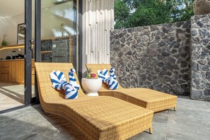 A sunlit outdoor seating area with wicker lounge chairs, a white table with a vase and fruit, and a stone wall backdrop. - Tropical 3BR Villa in Ubud w Pool, Patio & Garden (Gianyar Regency)