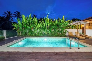 Ein Schwimmbecken mit einer gefliesten Kante, umgeben von üppigem Grün und einer Holzterrasse mit Liegestühlen und einem Sonnenschirm.