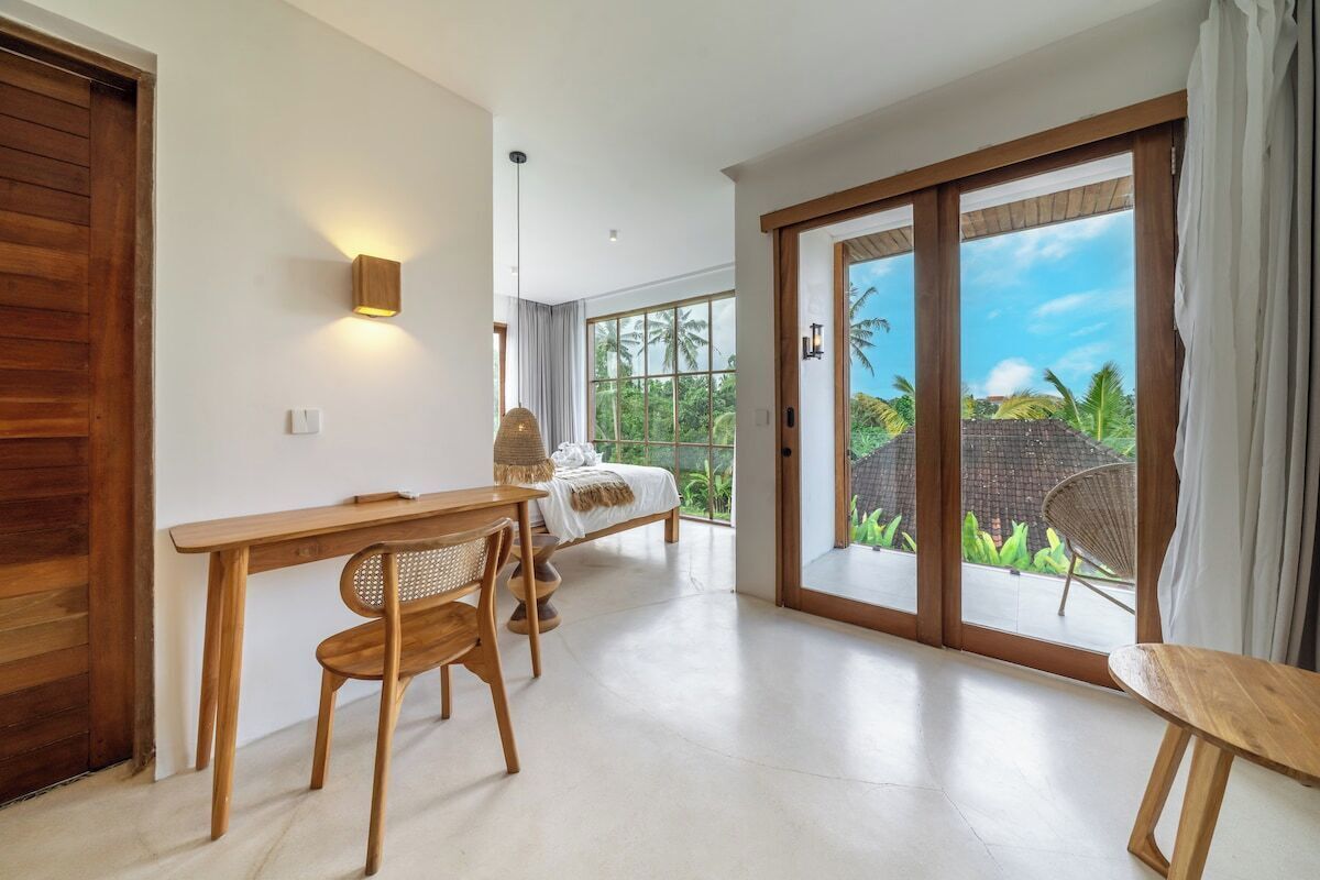 A room with a wooden desk, chairs, a bed, and a large sliding glass door leading to a garden.