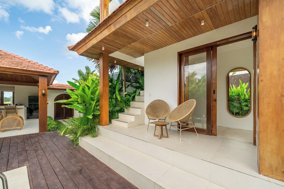 A covered outdoor area with a wooden deck, two wicker chairs, a staircase leading to a door, and lush greenery.