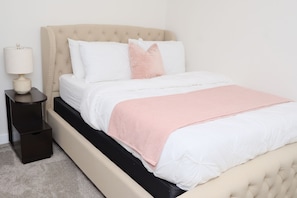 Un letto con una testiera beige, lenzuola bianche e una coperta rosa.
