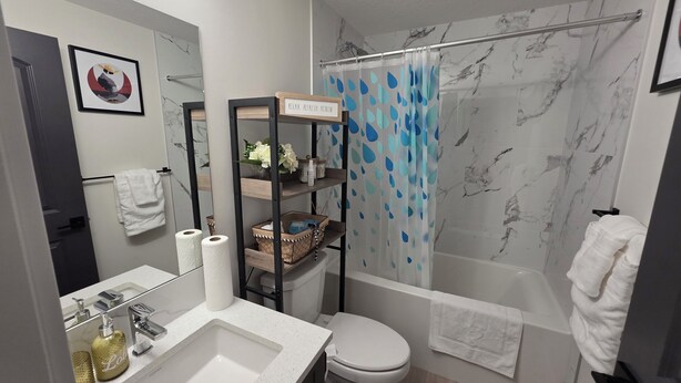 Un bany modern amb un lavabo, vàter i zona de dutxa de color blanc. Hi ha una prestatgeria amb articles de tocador i una fotografia emmarcada a la paret.