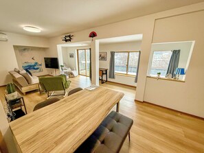 A modern living room with a wooden dining table, a sofa, and a green armchair. There is a television, a cactus, and a window with a view of the outside.