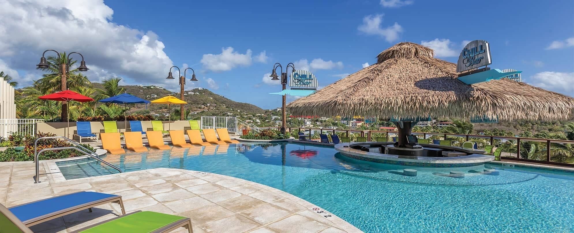 Zona de piscina con sillas de descanso, una estructura de techo de paja y un letrero que dice 'Chill Bar'.