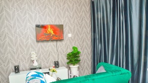 A hotel room with a modern design, featuring a large TV mounted on the wall, a comfortable green sofa, a white console table with a vase of flowers, and a potted plant. - Alasiri Apartments Utawala (Nairobi)