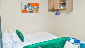 A single bed with white bedding, a green accent pillow, and a blue and white blanket. A wooden cabinet with a blue hanger is mounted on the wall. - Alasiri Apartments Utawala (Nairobi)