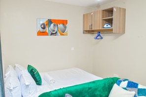 Cama de solteiro com roupa de cama branca, travesseiro de destaque verde e manta azul e branca. Armário de madeira com cabide azul fixado na parede.