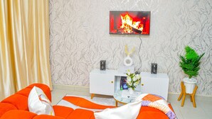 A living room with a sofa, a TV displaying a fire, a white console table with speakers, and a potted plant. - Alasiri Apartments Utawala (Nairobi)