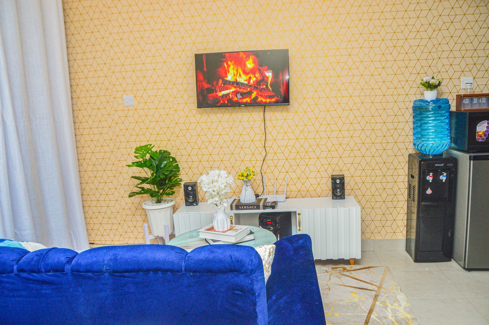 Una sala de estar con un sofá azul, una mesa blanca con un altavoz, una planta y un cuadro en la pared con una imagen de un fuego.