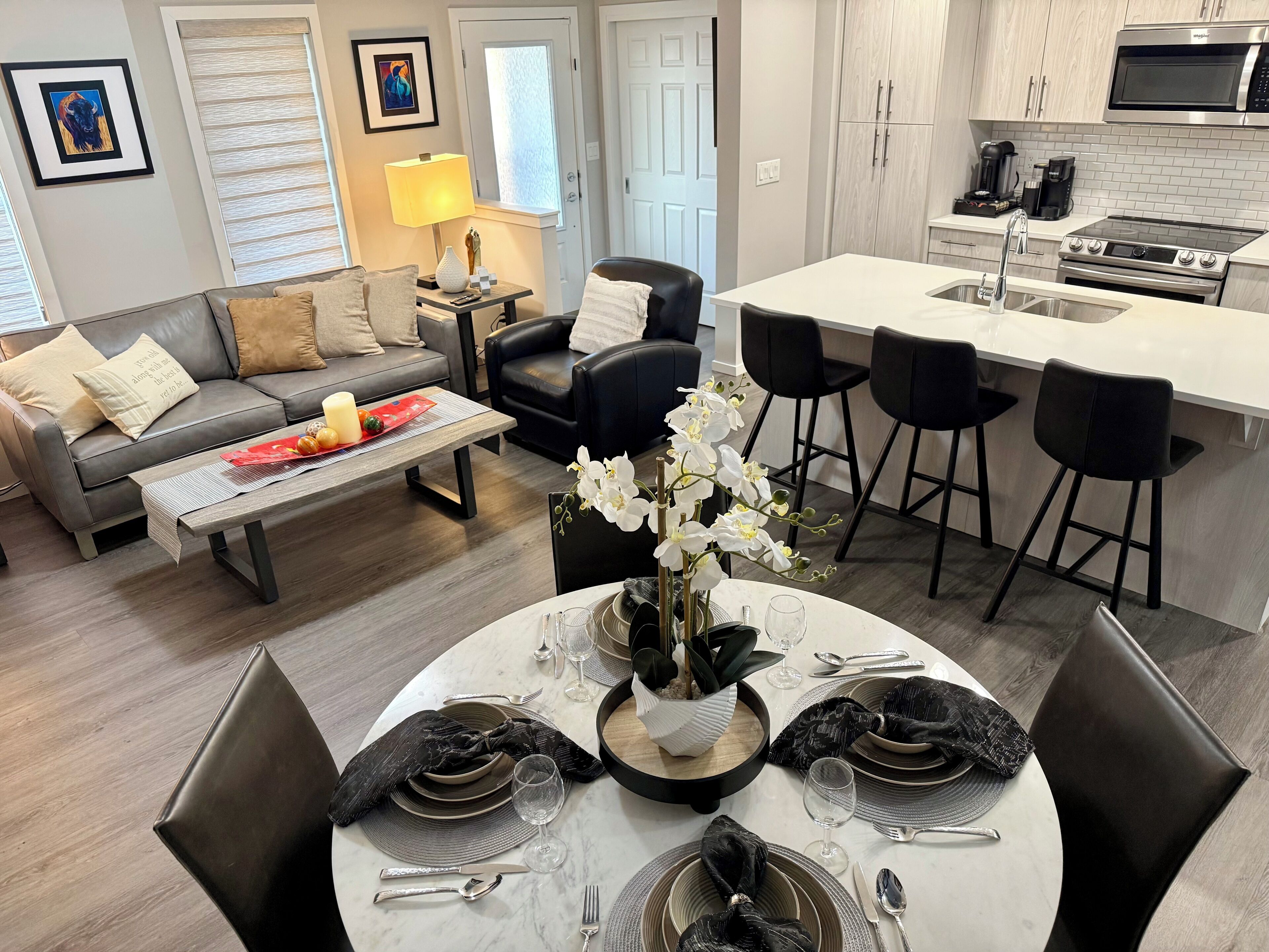 Un salon moderne avec un canapé gris, une table basse et un coin repas aménagé pour quatre personnes, avec une nappe blanche et des serviettes noires.