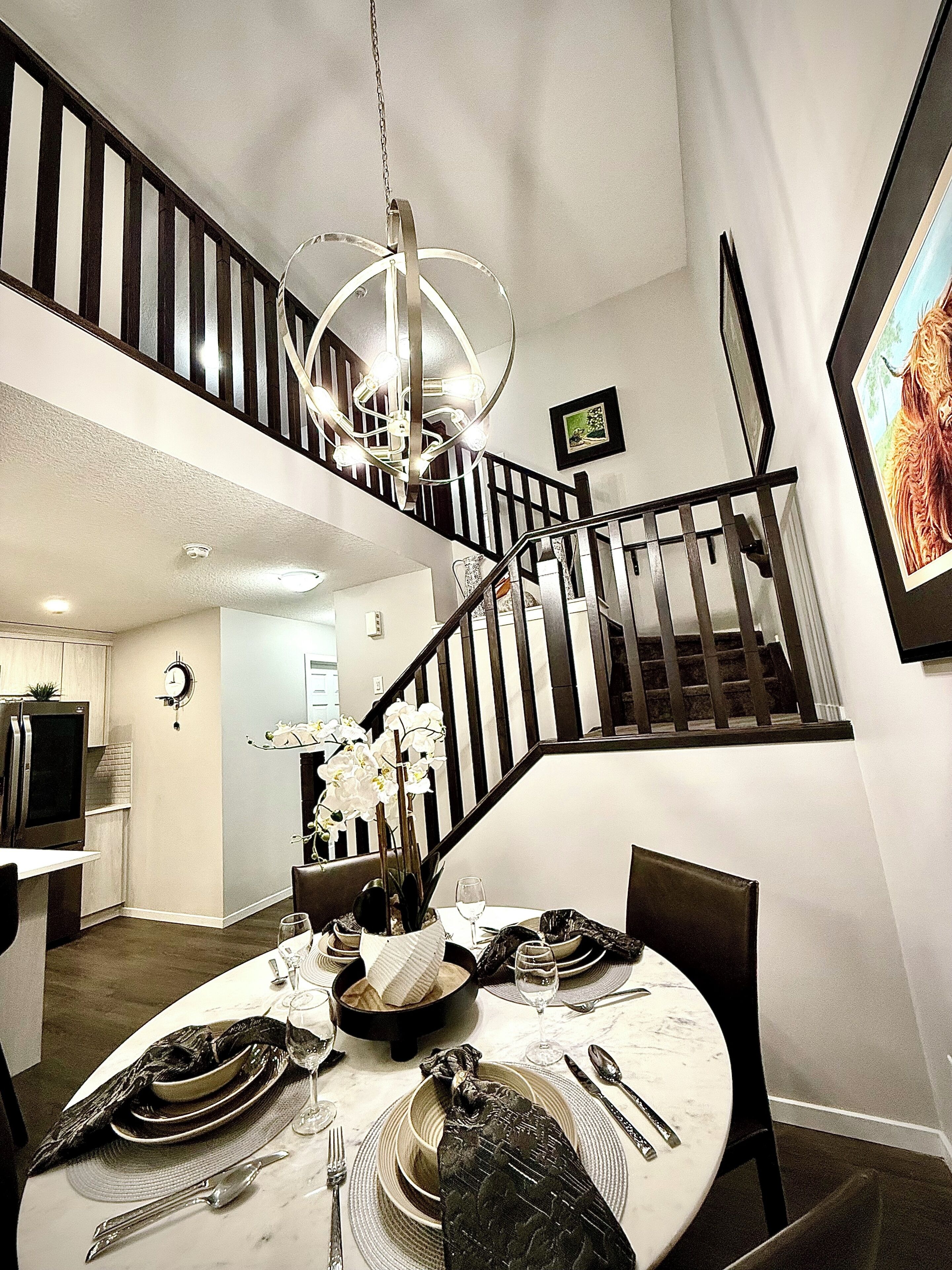 A dining area with a round table set for four, a chandelier, and a staircase leading to an upper level.