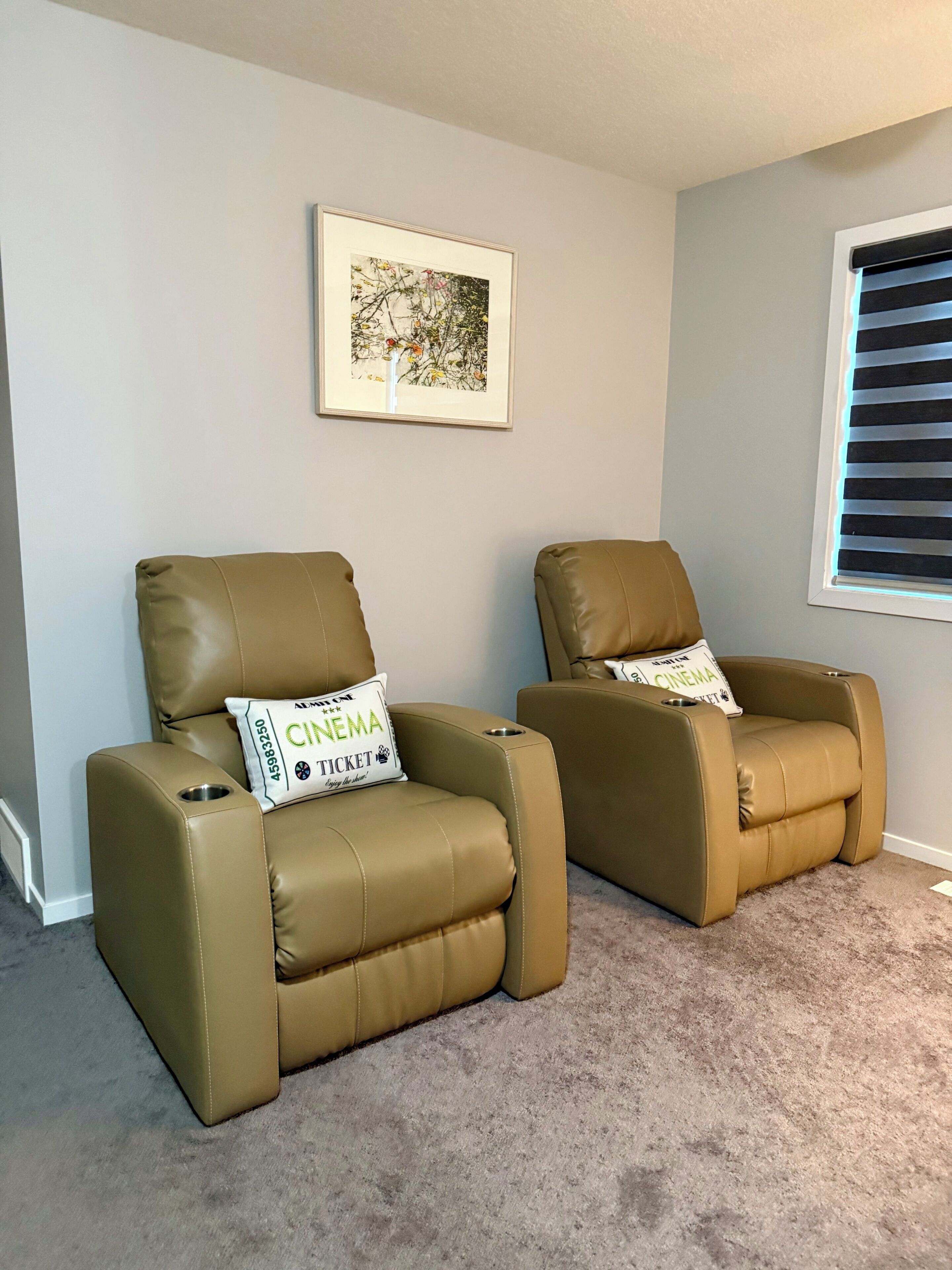 Twee beige leren verstelbare fauteuils met 'CINEMA' kussens, een ingelijste foto aan de muur en een raam met zonwering.