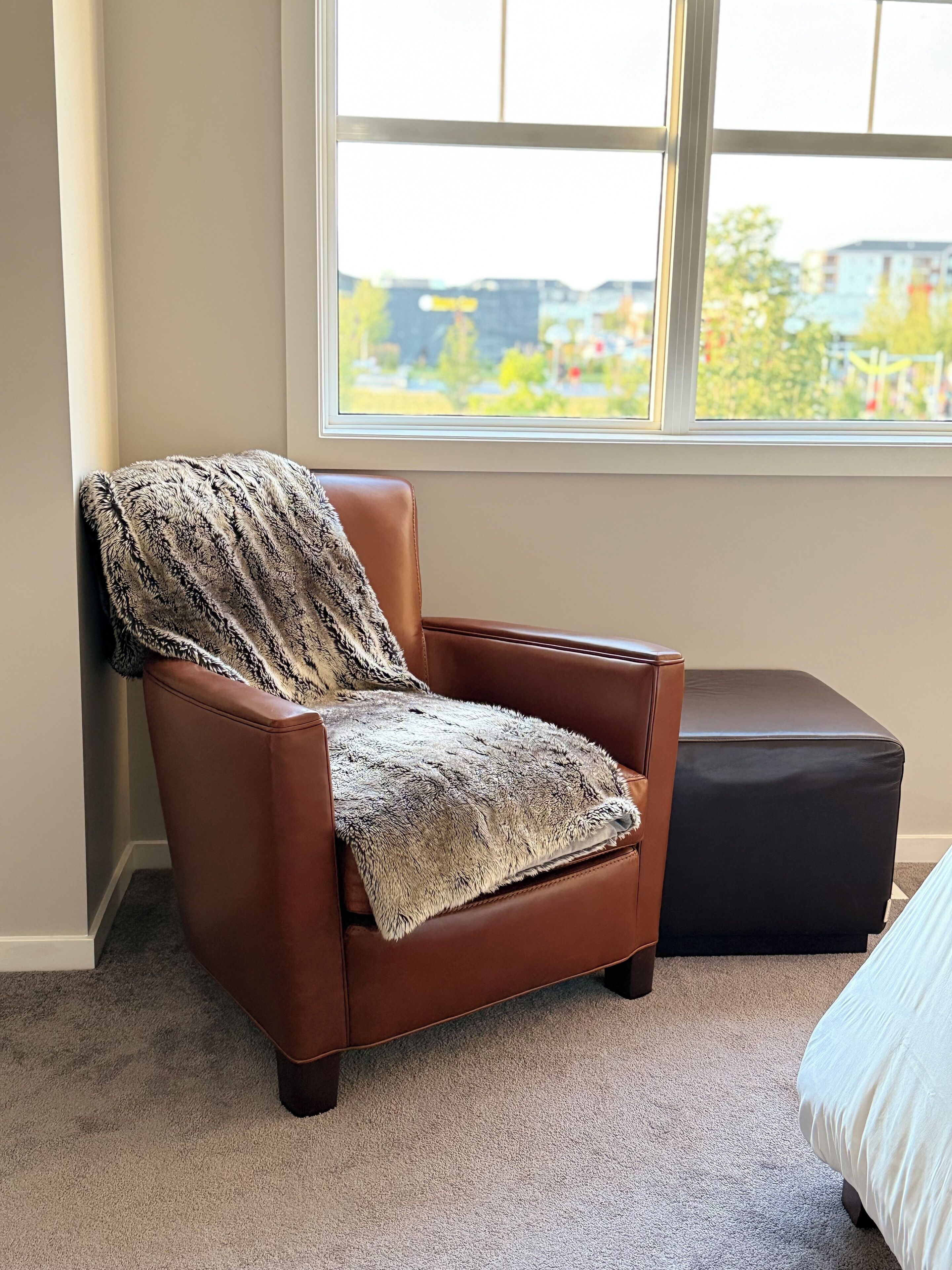 A brown leather armchair with a patterned throw blanket, positioned next to a black ottoman and a bed with a white duvet, in a room with a large window offering a view of a building and greenery.