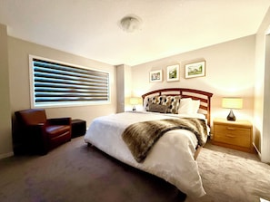 A bedroom with a bed, a chair, a nightstand with a lamp, a window with blinds, and framed pictures on the wall. - Free Car garage|3BR|YYC|Banff|Canmore|Winsport|Stampede (Calgary)