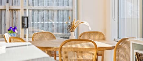 Un área de comedor con una mesa de madera y sillas tejidas, un florero con plantas secas, y un ventanal con vista a una cerca de madera.