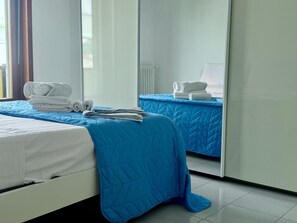 Et hotelværelse med to senge, hvoraf den ene har et blåt tæppe og den anden hvide sengetøj. Der ligger foldede håndklæder på hver seng.