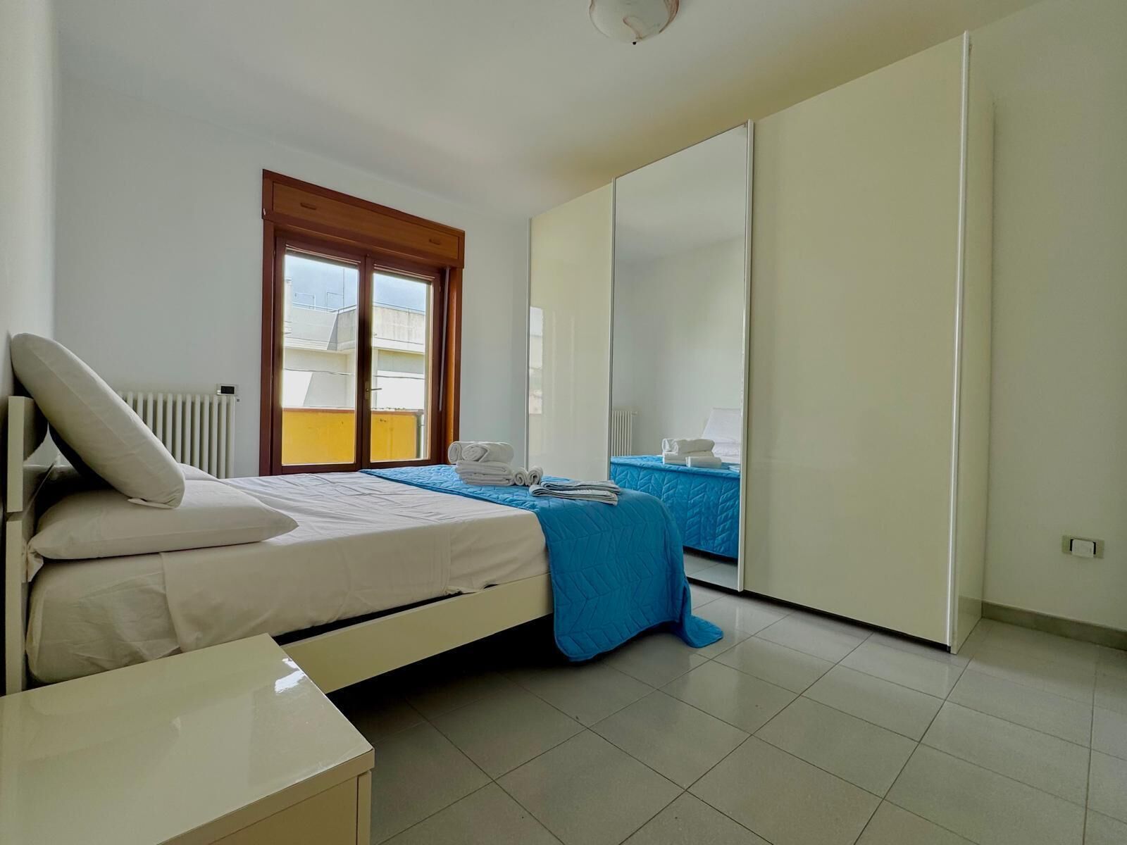 A bedroom with a bed, a bedside table, a window, a wardrobe, and a view of the outside.