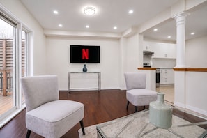 A modern living room with two armchairs, a glass coffee table, a TV mounted on the wall, and a kitchen area visible through an open doorway. - Luxe Chicopee Townhome: Ravine Forrest, Sleeps 8 (Kitchener)