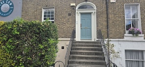 A brick building with a blue door, a staircase, and a hedge in the foreground.