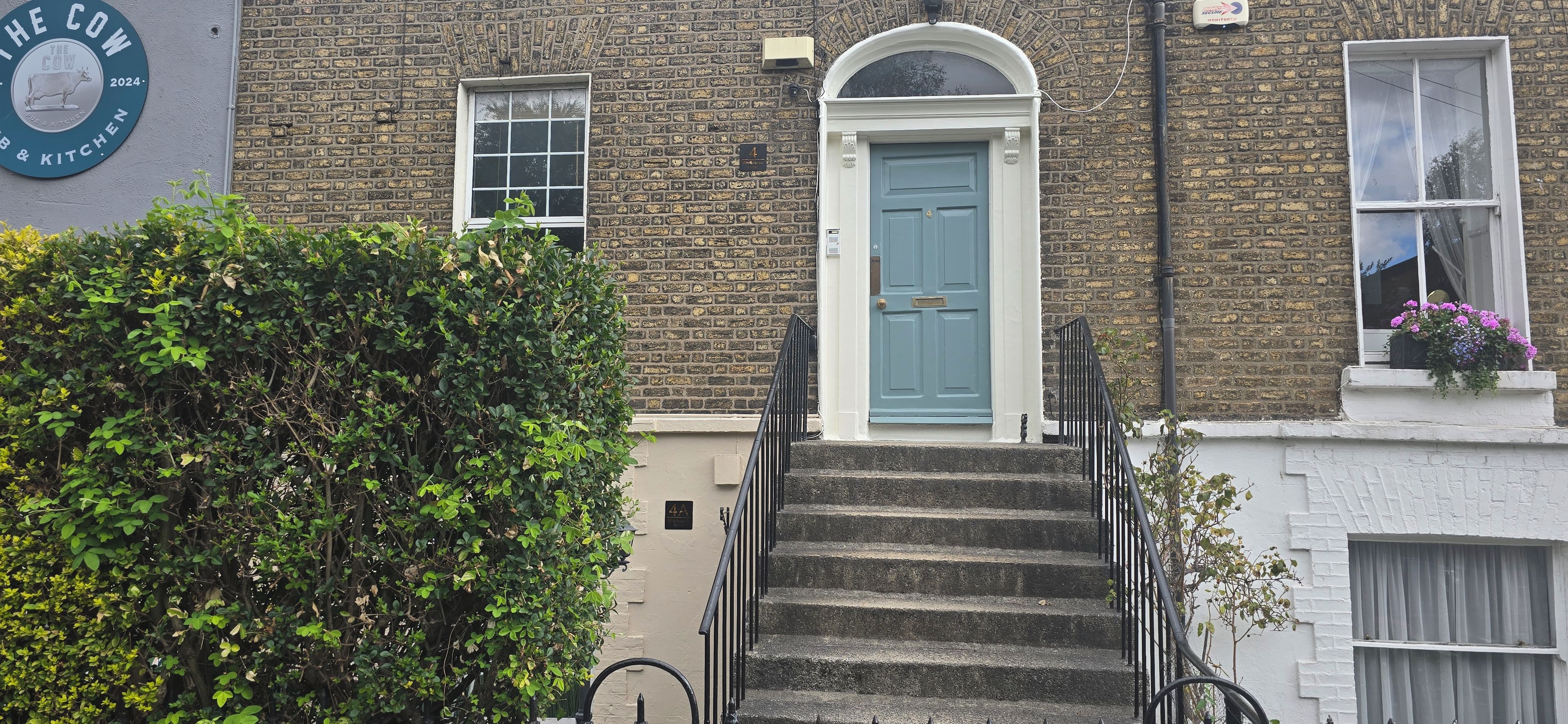 A brick building with a blue door, a staircase, and a hedge in the foreground.