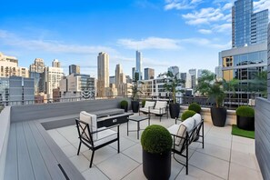 A rooftop terrace with outdoor furniture, potted plants, and a city skyline view.