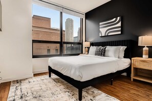 Ein modernes Schlafzimmer mit großem Fenster, einem Bett mit weißer Bettwäsche, einem schwarzen Kopfteil und Blick auf die Stadt.