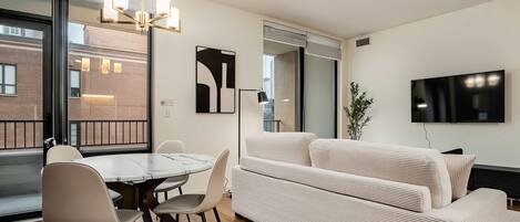 A modern living room with a dining area, a sofa set, a flat-screen TV, and a large window with a view of the city.