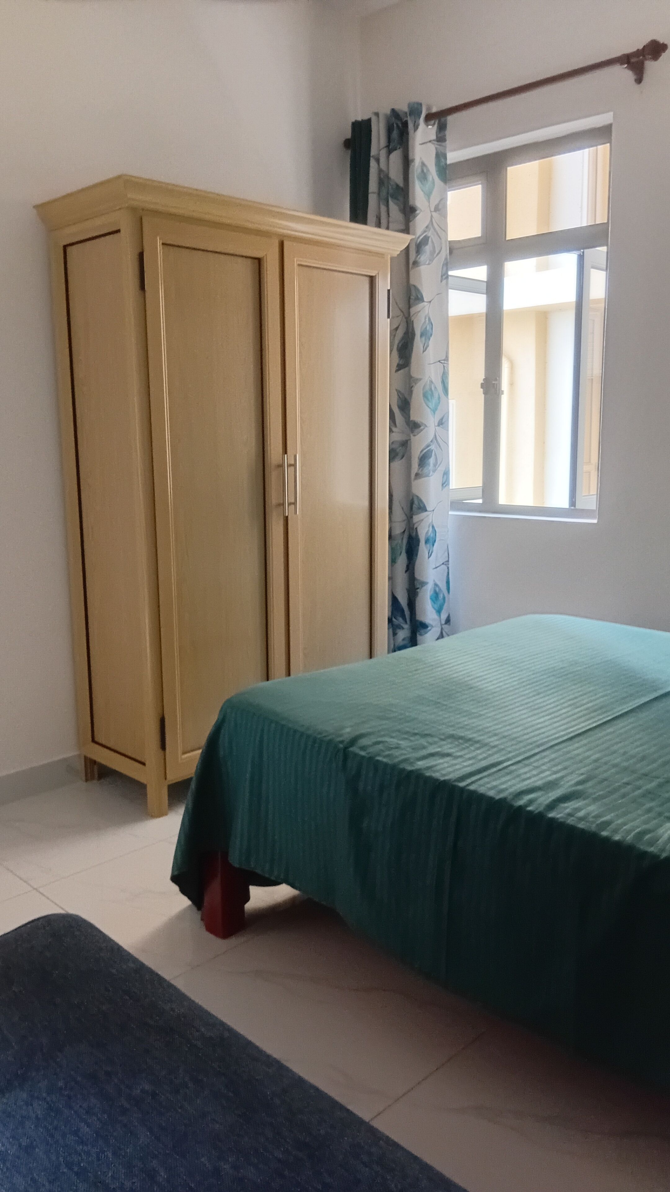 A bedroom with a bed, a wardrobe, a window with curtains, and a tiled floor.