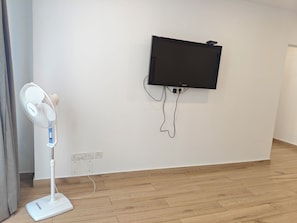 A room with a standing fan, a mounted television, and a power outlet on the wall.