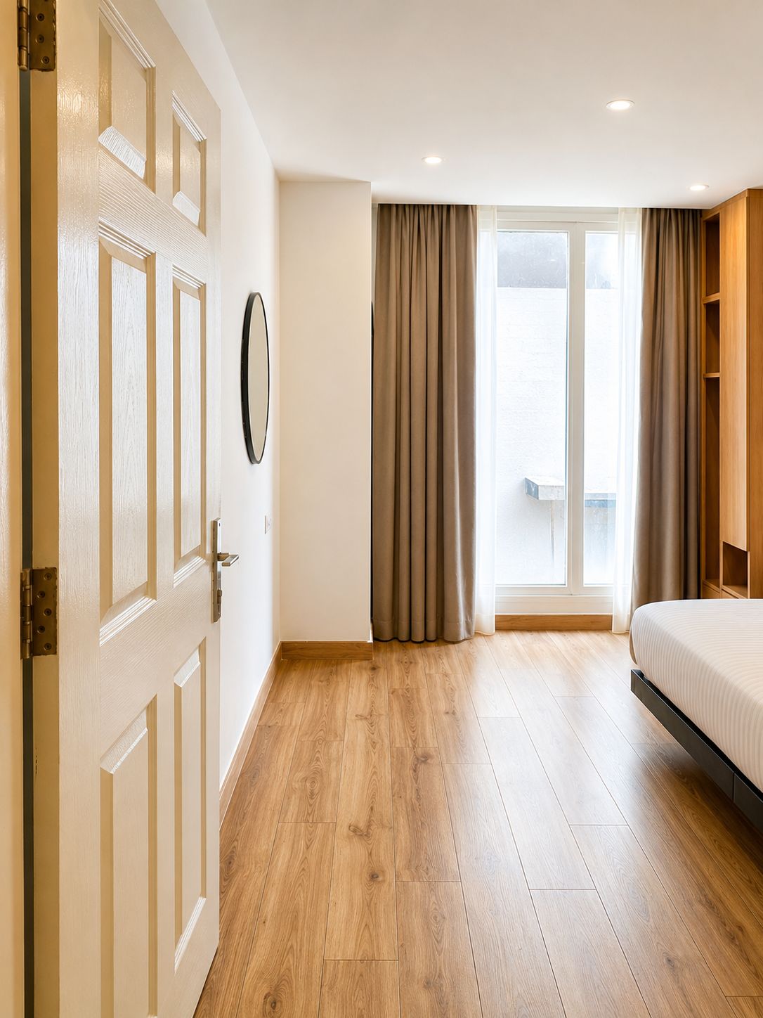 A hotel room with a wooden floor, a bed, a door, a window with curtains, and a wall-mounted mirror.