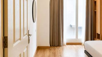 A hotel room with a wooden floor, a bed, a door, a window with curtains, and a wall-mounted mirror.