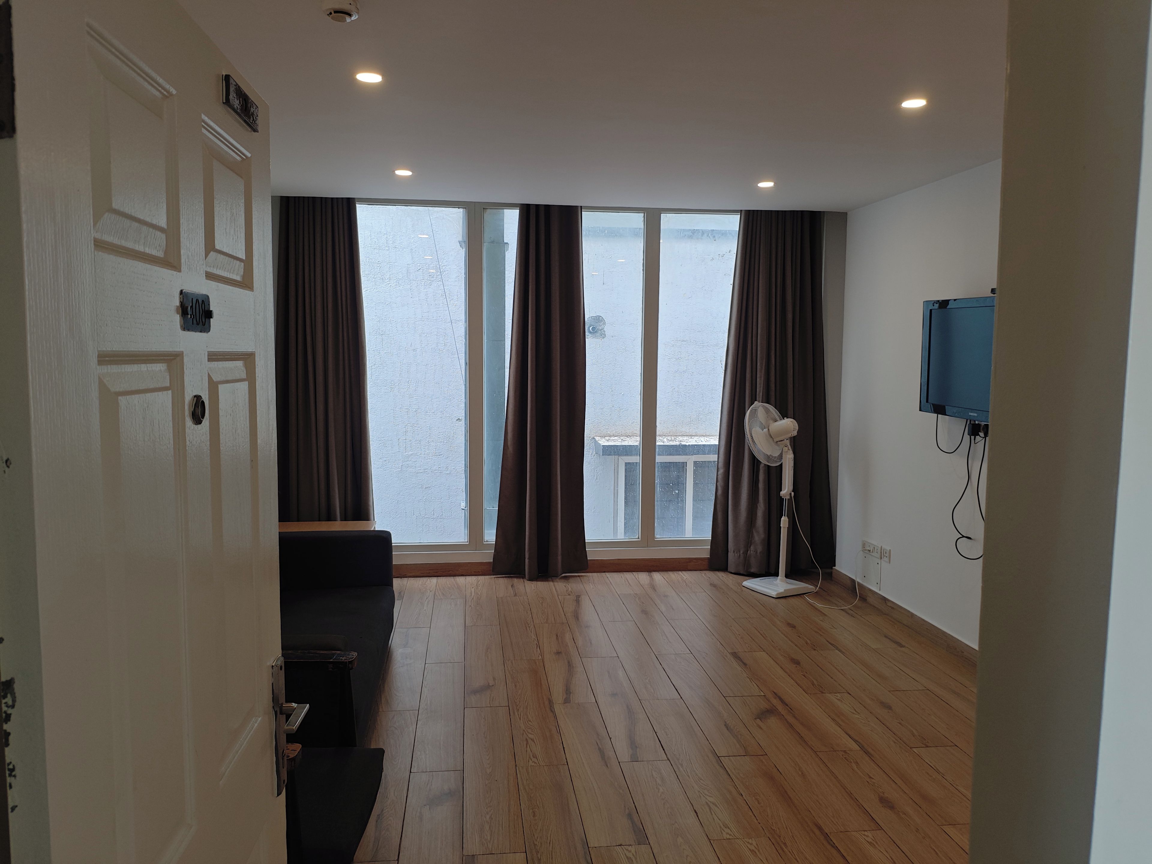 A room with a wooden floor, a black sofa, a standing fan, a TV mounted on the wall, and a large window with curtains.