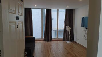 A room with a wooden floor, a black sofa, a standing fan, a TV mounted on the wall, and a large window with curtains.