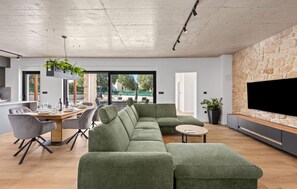 A modern living room with a sectional sofa, dining table, and chairs. There is a flat-screen TV mounted on a stone wall and a potted plant on the floor. - Gorgeous home in Dubrava Kod Sibenika with sauna (Dubrava Kod Sibenika)