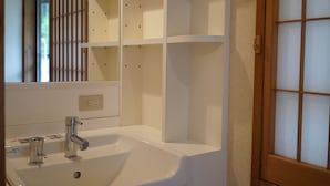 Casa de banho com lavatório branco, duas torneiras e espelho. Há prateleiras acima do lavatório e uma porta de madeira com painéis de vidro.