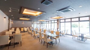 Un amplio comedor interior con múltiples mesas y sillas, ventanas grandes que permiten la entrada de luz natural y conductos de ventilación en el techo.
