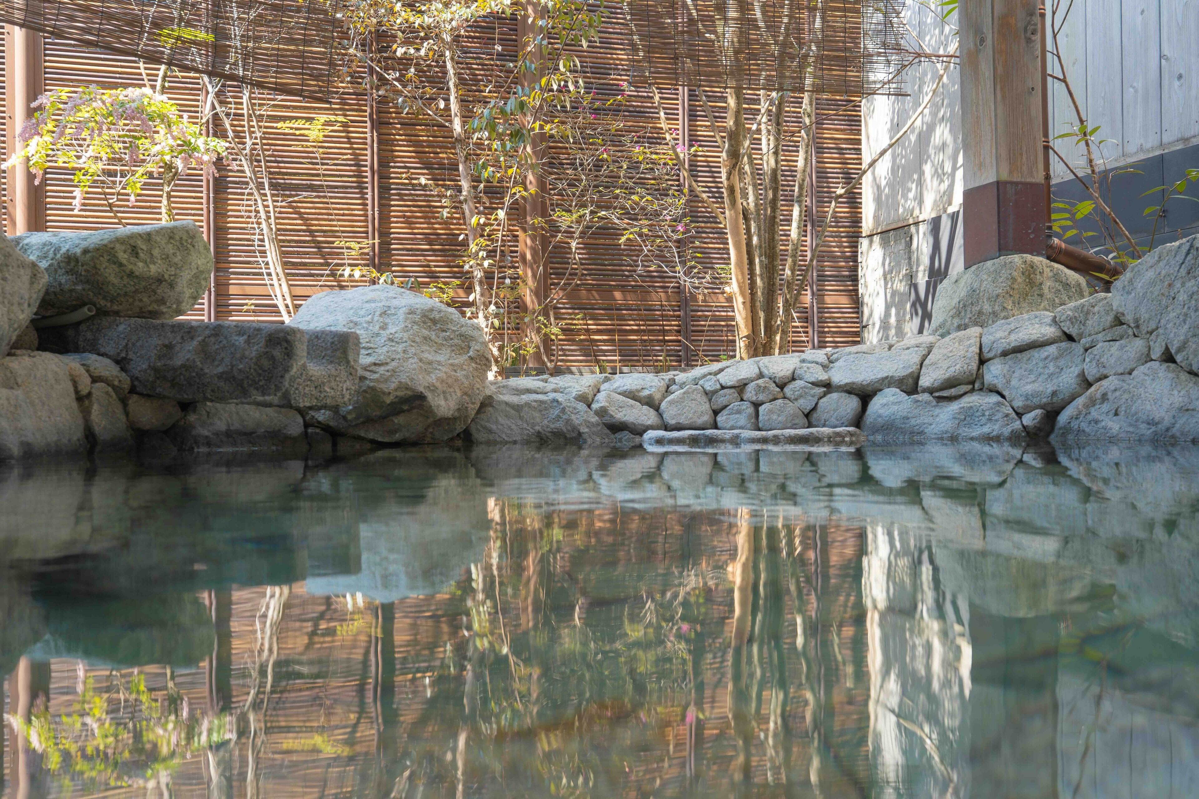 A tranquil outdoor pool area with clear water, surrounded by rocks and bamboo walls.