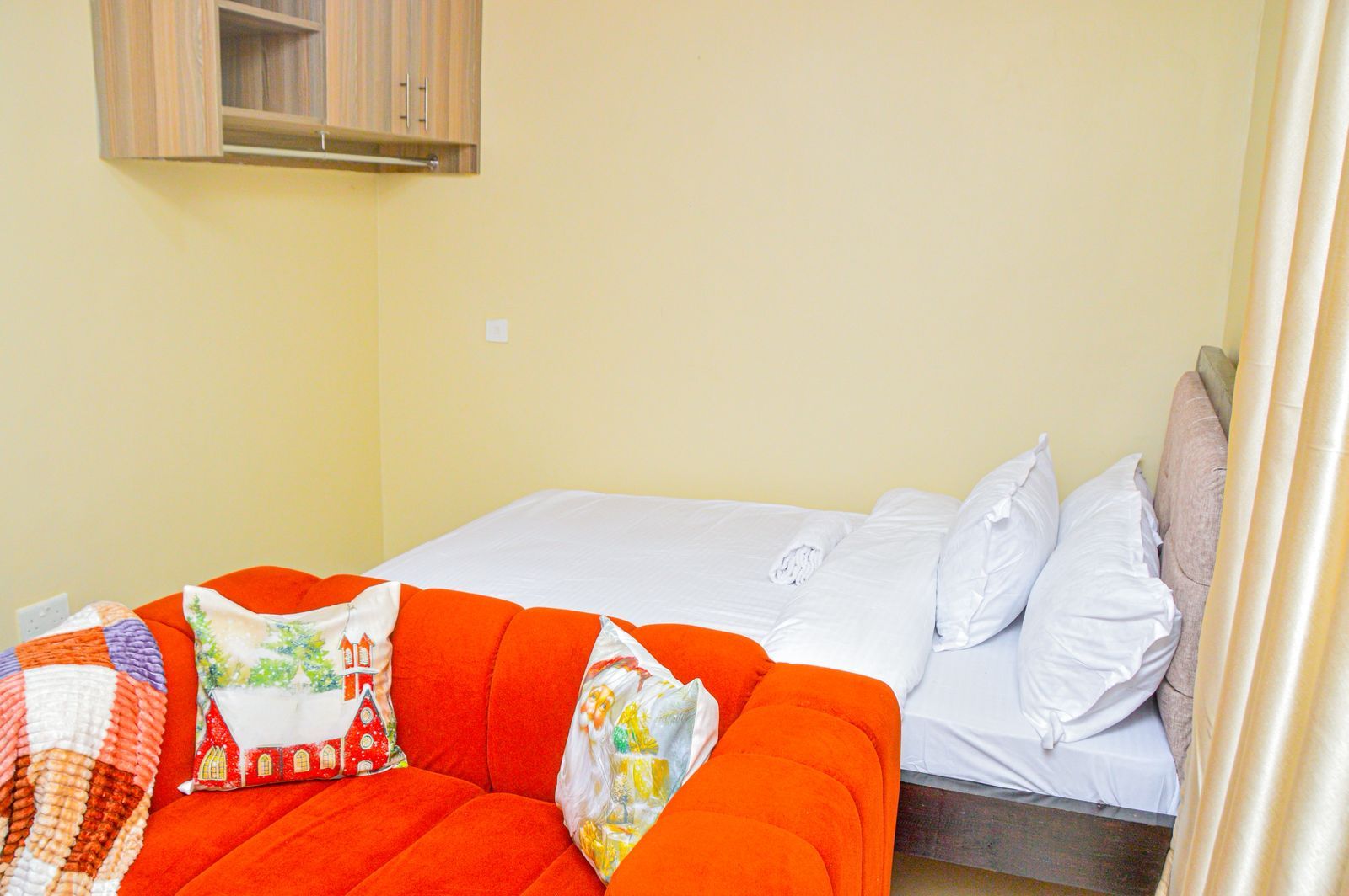 A bed with white bedding, a red sofa with patterned cushions, and a wooden cabinet above the bed.