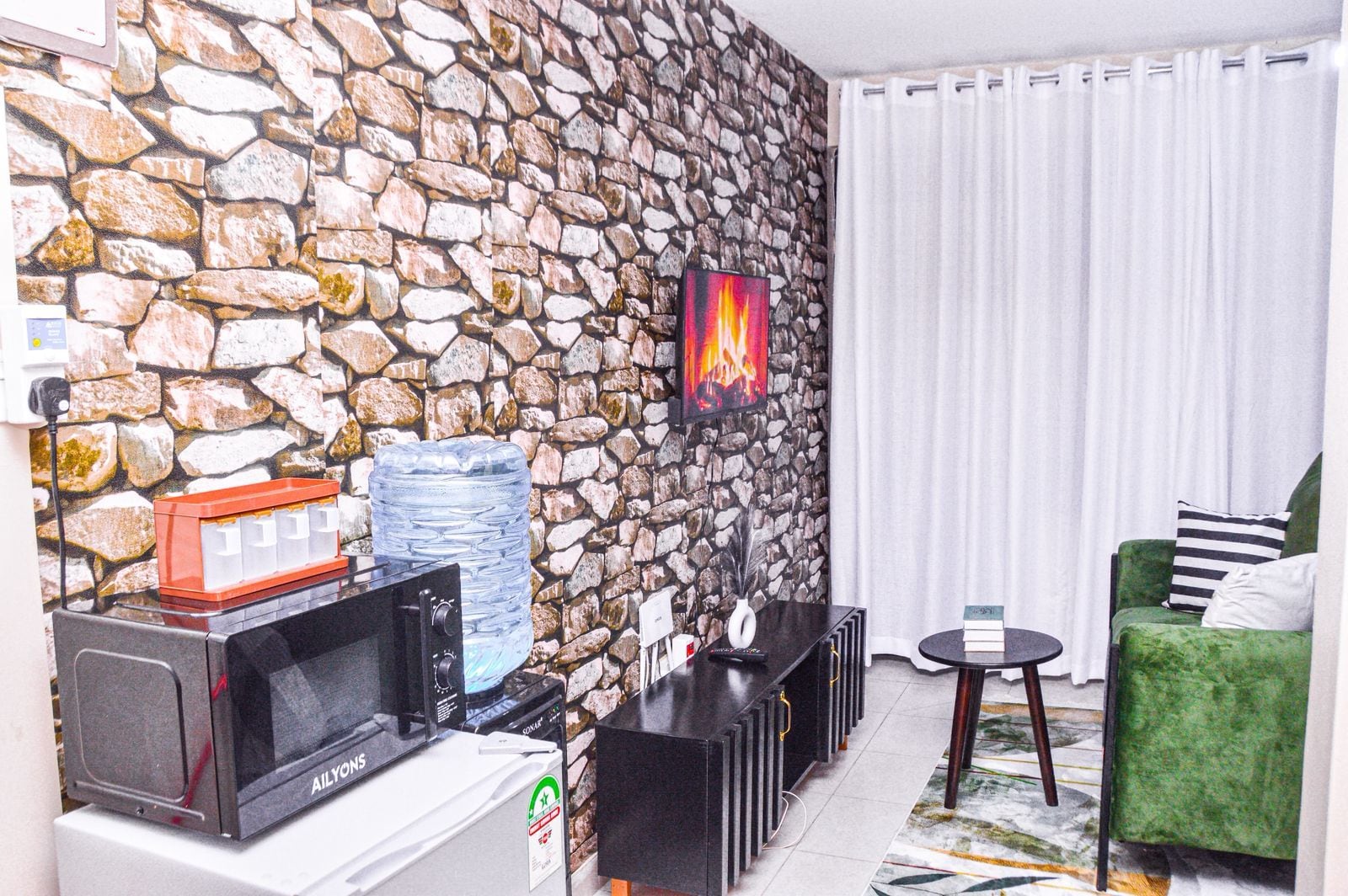 Sala de estar com parede de pedra, televisão de tela plana, dispensador de água, micro-ondas, mesa pequena e poltrona verde.