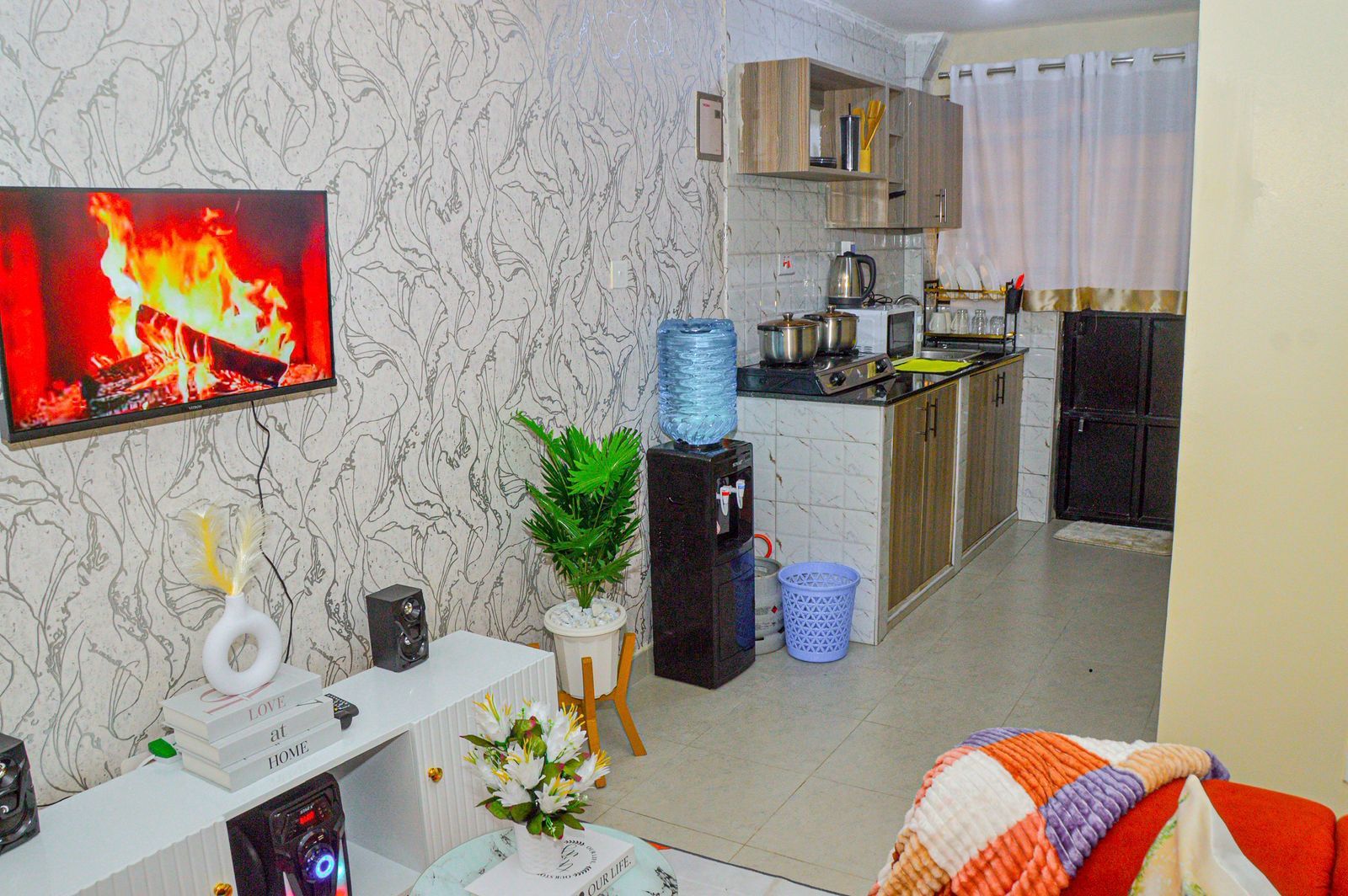 Una sala de estar con un televisor mostrando un fuego, un mueble bajo blanco con objetos decorativos, una zona de cocina con armarios y un dispensador de agua.