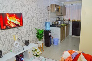 Una sala de estar con un televisor mostrando un fuego, un mueble bajo blanco con objetos decorativos, una zona de cocina con armarios y un dispensador de agua.