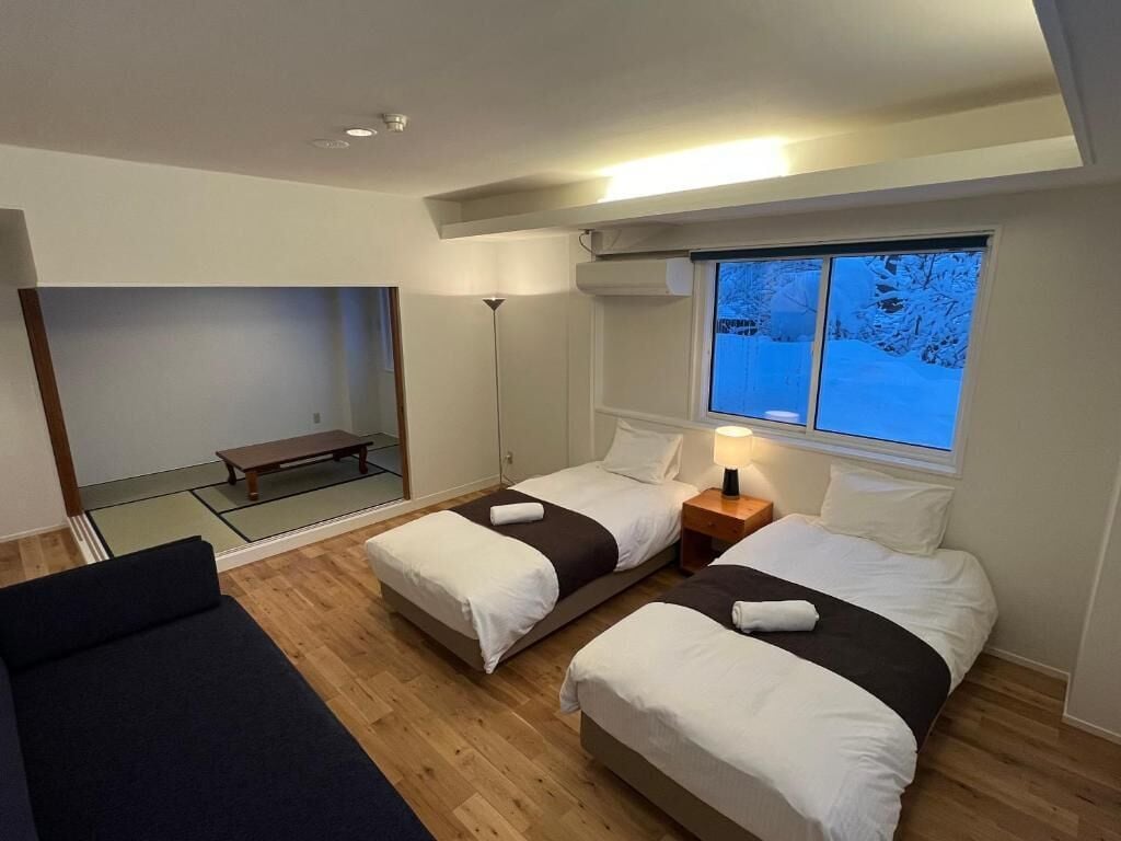 Een hotelkamer met twee bedden, een bankje, een staande lamp en een raam met uitzicht op bomen.