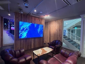 Habitación con una gran televisión de pantalla plana montada en una pared de madera, sillones de cuero, una mesa de centro de mármol y una puerta de cristal que da a una escalera.