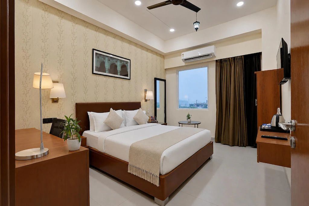 A hotel room with a large bed, a desk with a lamp, a plant, an air conditioning unit, a television, and a view of the city through the window.
