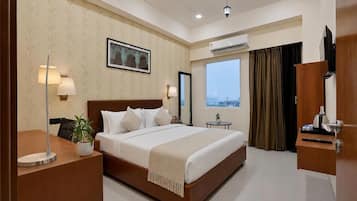 A hotel room with a large bed, a desk with a lamp, a plant, an air conditioning unit, a television, and a view of the city through the window.