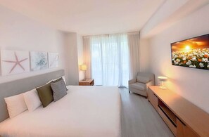 Ein Hotelzimmer mit einem großen Bett, einem Sessel, einem Schreibtisch und einem Fernseher an der Wand.