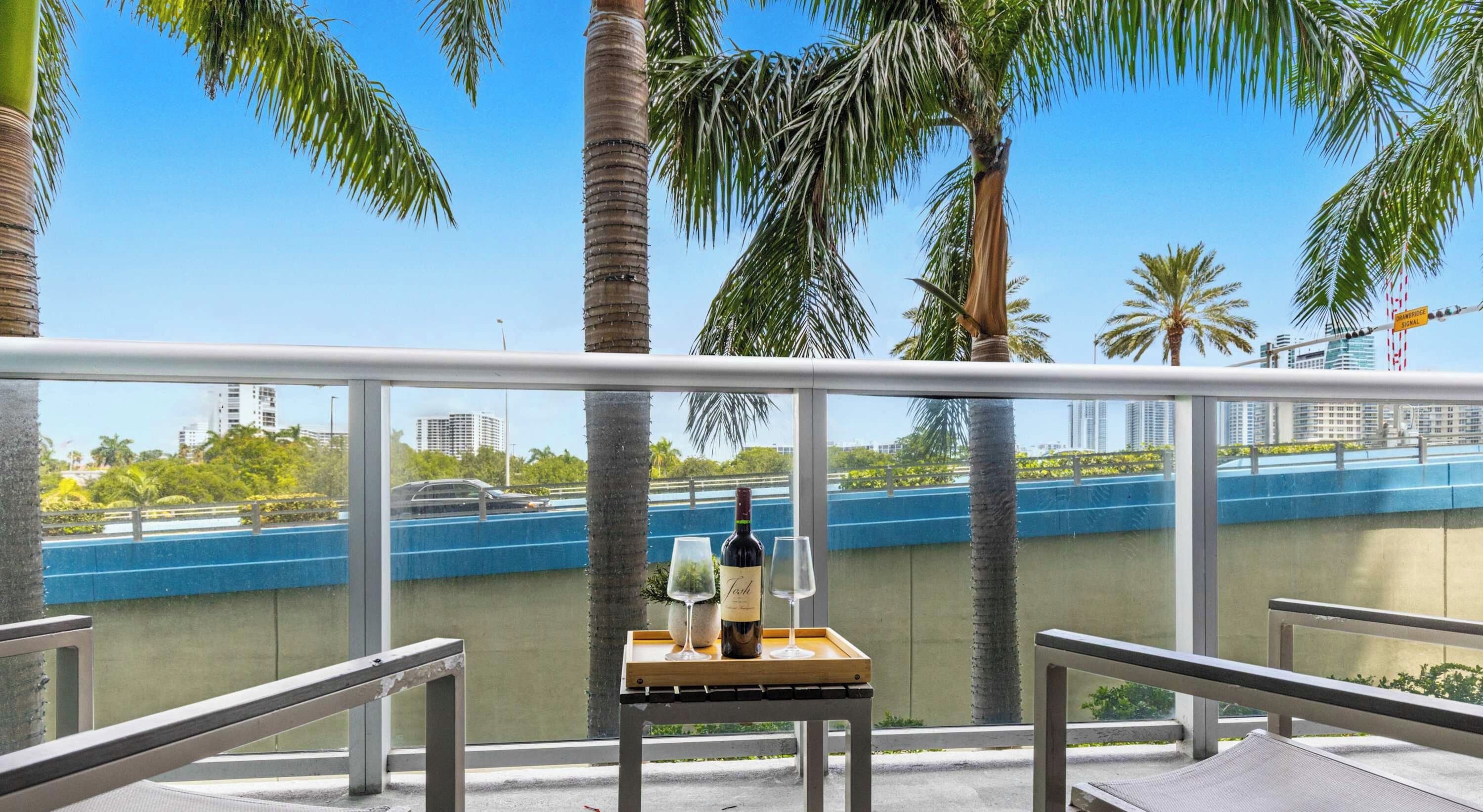 A balcony with a table set for two, overlooking a blue waterfront and palm trees.